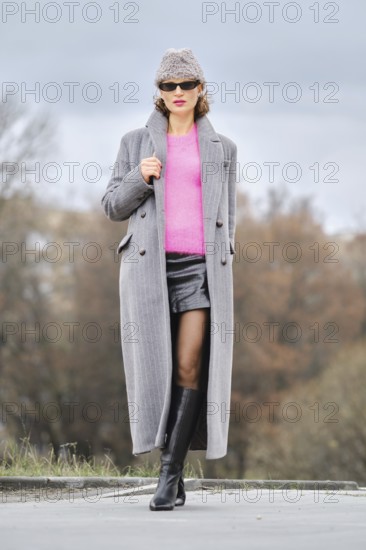 A stylish woman walks down a city street, wearing a pink sweater, unbuttoned long coat, and leather