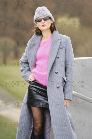 A woman stands playfully in city park in a stylish autumn outfit, showcasing urban street style.