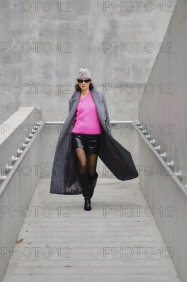 A fashionable woman in sunglasses rushing down a sleek grey walkway, wearing a mini leather skirt,