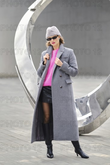A woman in a long gray coat and pink sweater posing near a large modern sculpture. She wears black