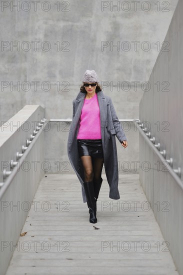 A stylish woman walks down a sleek urban walkway, dressed in a stylish gray coat over a bright pink