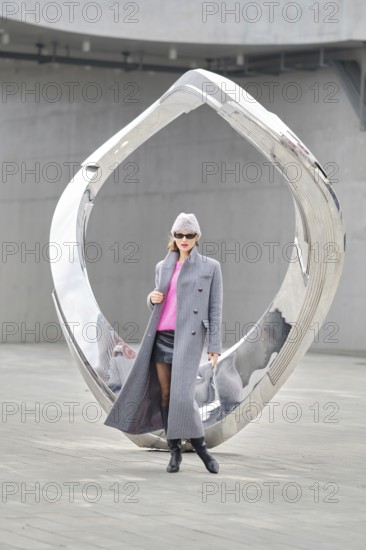 A stylish woman wears a grey coat and pink sweater while posing by a sleek, reflective sculpture in