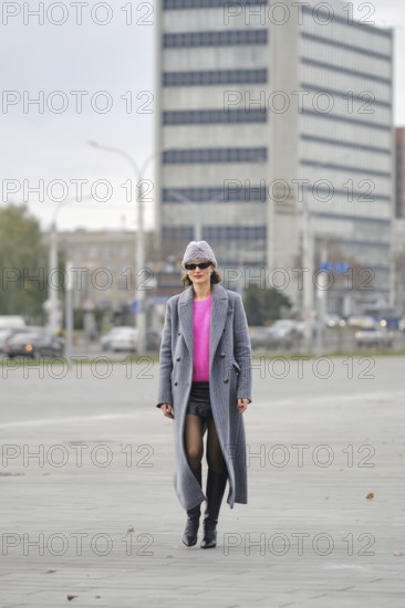 A stylish woman walks through an urban setting wearing a gray long coat and a bright pink sweater