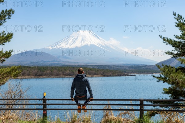 Young man sitting on railings next to a road and looking across the lake to Mt Fuji volcano, Motosu