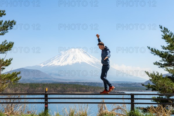 Young man balancing on railings next to a road, view across the lake to Mt Fuji volcano, Motosu