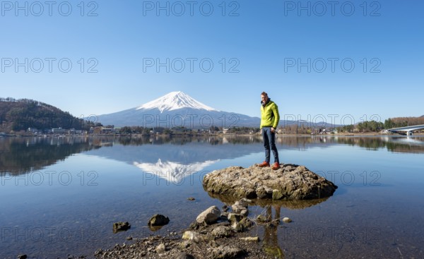 Young man standing on a rock in water, volcano Mt. Fuji is reflected in Lake Kawaguchi, Yamanashi