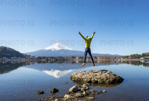 Young man standing on a rock in water, jumping into the air, volcano Mt. Fuji is reflected in Lake