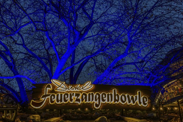Sales stand for Feuerzangenbowle, above a deciduous tree illuminated with blue light,