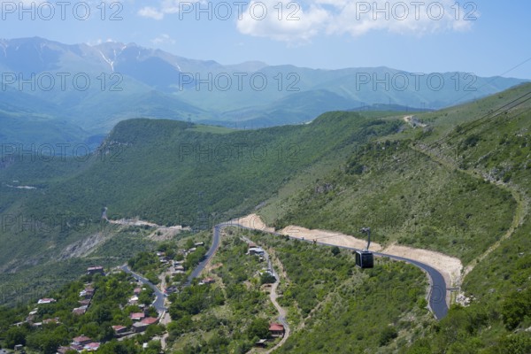 A winding road runs through green hills and passes a small village, with views of mountains, Tatev