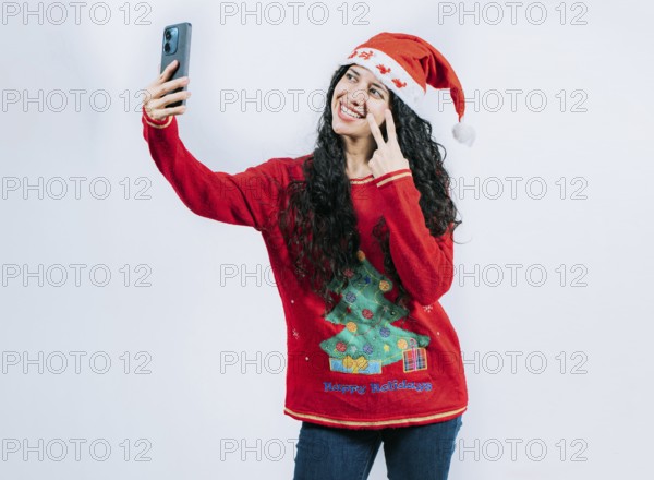 Funny girl in Christmas hat and sweater taking selfie, isolated. Happy young latin woman in