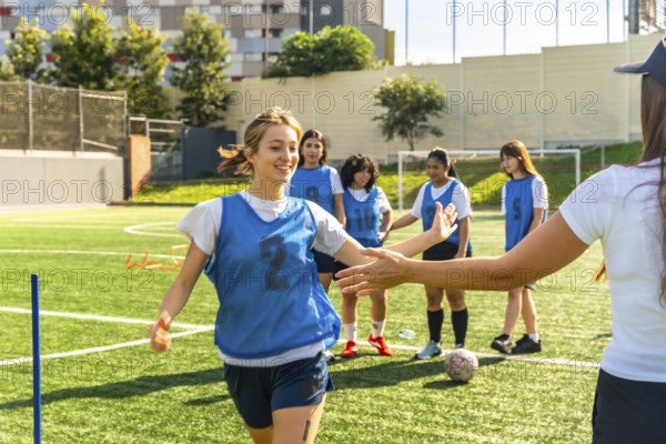 Young female soccer player in a blue bib high fiving her coach on a green artificial turf field,