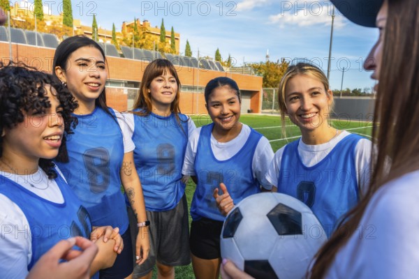 Diverse women football players smiling and listening, holding a soccer ball on a green artificial