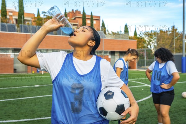 Young woman soccer player in a blue bib holding a football, refreshing herself by drinking a sports