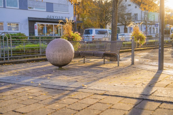 Public bench and large sphere on paved square, autumn trees and buildings in the background,