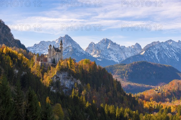Neuschwanstein Castle in a forest of colorful autumn trees, against snow-covered mountains and blue