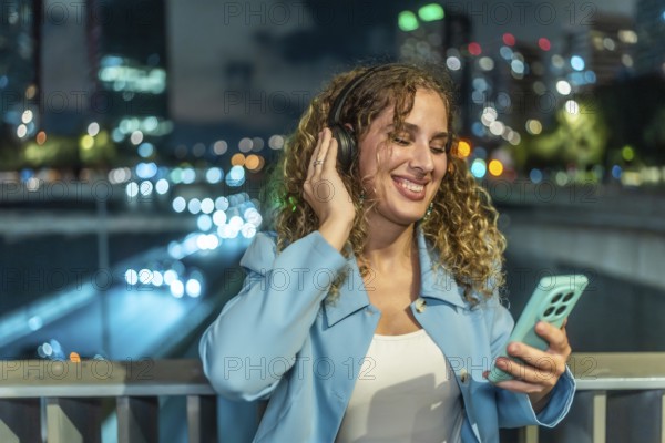Woman with headphones and smartphone enjoying music while smiling cheerfully, standing on a bridge