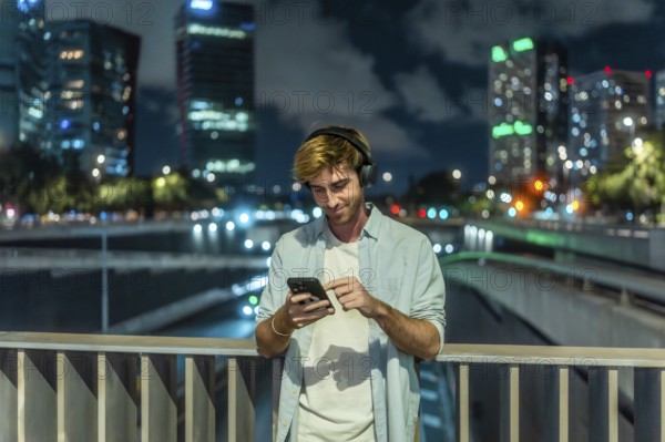 Young man wearing headphones and casual clothing standing on a city bridge at night, smiling and
