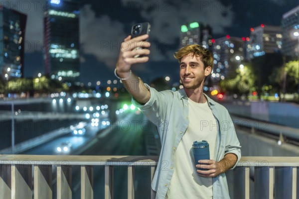 Young man smiling and taking a selfie with a smartphone, holding a reusable coffee cup on an