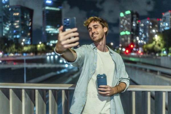 Young man smiling and taking a selfie with a smartphone while holding a reusable coffee cup,