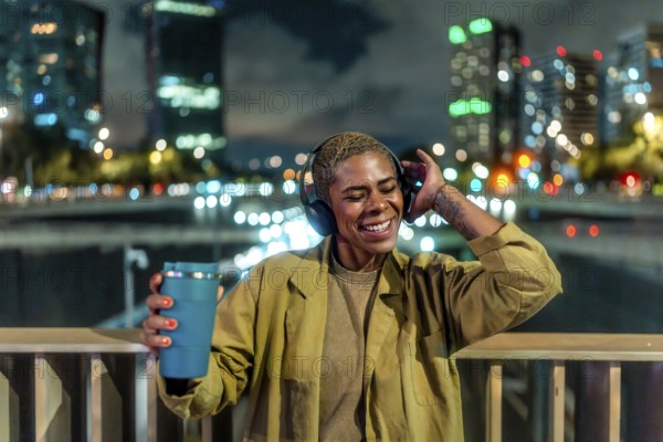Happy young black woman standing on an urban bridge at night, enjoying music through headphones and