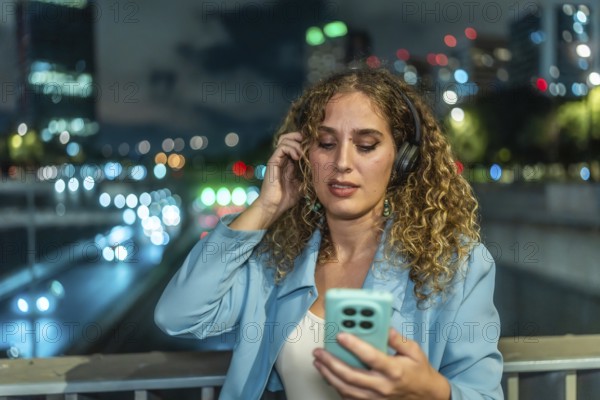 Young woman with curly hair wearing headphones and looking at her smartphone while enjoying music
