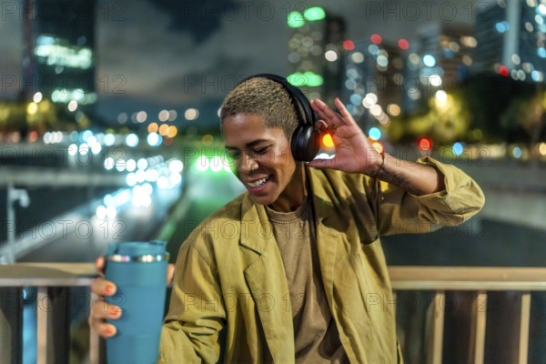Woman listening to music with headphones and holding a travel mug, smiling brightly while standing