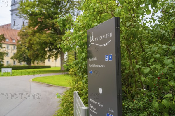 A sign with the reference to Zwiefalten stands against a background of green plants and a building,