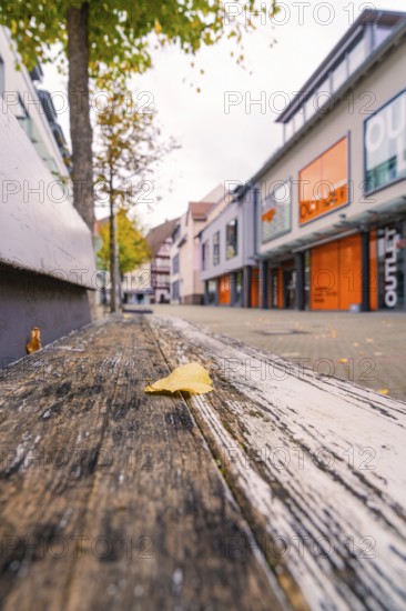 A quiet shopping street with modern buildings, autumn leaves and a wooden bench in the foreground,