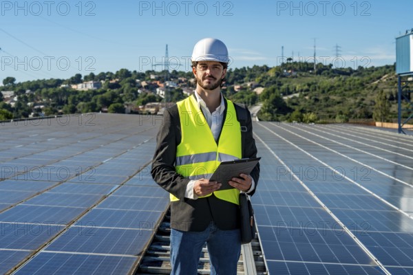 Engineer in hard hat and safety vest standing on a rooftop of solar panels holding a clipboard,