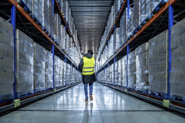 Worker in high visibility clothing walking down a long aisle between rows of elevated racks packed