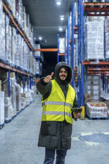 Warehouse worker in warm clothing and safety vest holding a barcode scanner and giving thumbs up