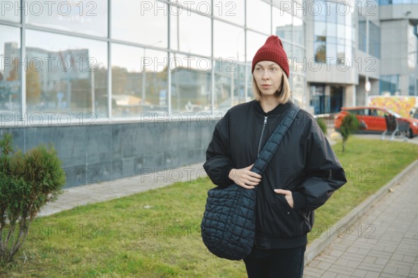 A woman wearing a black jacket and red beanie walks along a modern glass building. She holds a