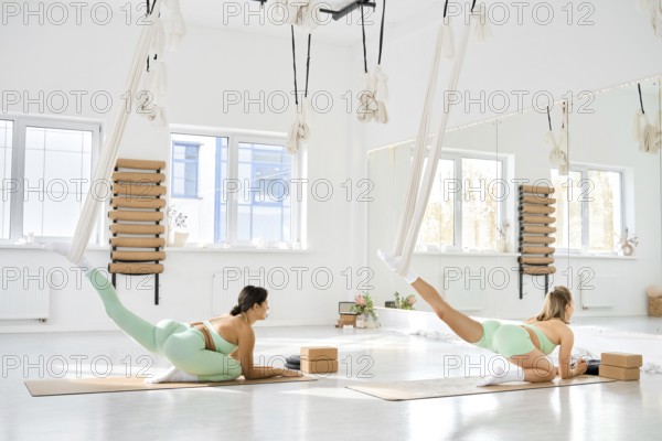 Two individuals are practicing fitness stretching in a well-lit studio. They use hammocks for