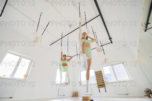 Two individuals engage in a fitness stretching routine using a hammock in a bright, spacious studio