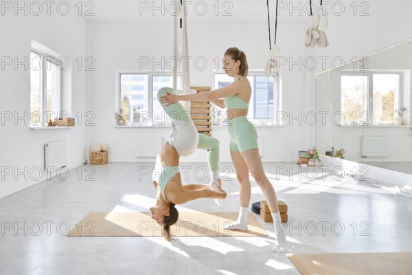 Two women engage in fitness stretching using a hammock in a spacious, well-lit studio. The