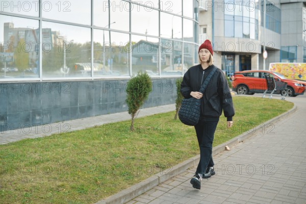 A woman strolls along a wide walkway surrounded by a mix of urban buildings and greenery. She wears
