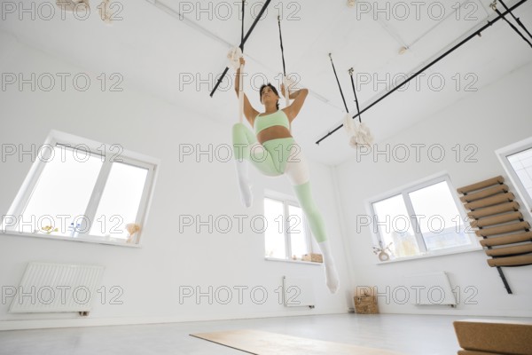 A woman in green activewear performs stretching exercises with the support of a hammock. She is in