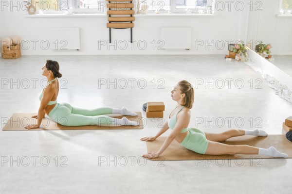 Two women are engaged in yoga practice on mats in a sunlit studio. They are positioned in deep