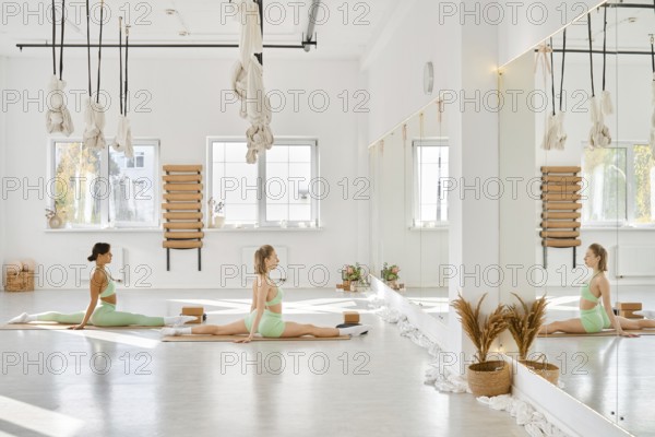 In a spacious yoga studio filled with natural light, two women perform stretches on mats. The room