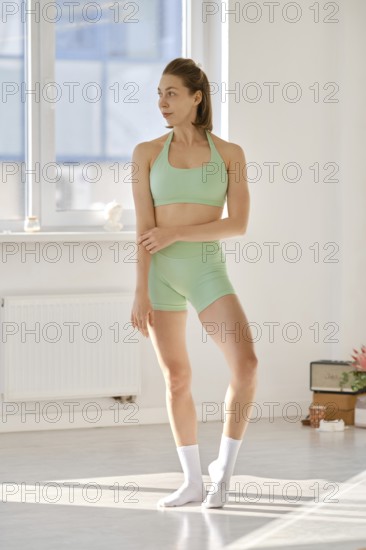 A woman in a green outfit stands gracefully in a well-lit yoga studio. She appears focused and