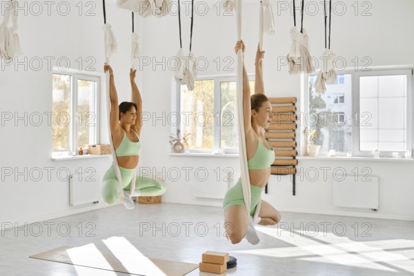 Two women practice fitness stretching with hammocks in a bright studio. They focus on flexibility
