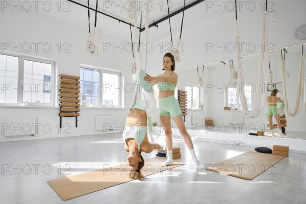 Two fitness enthusiasts are participating in a stretching session using a hammock in a bright, airy