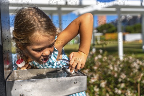 Little girl bending to drink cool water from a stainless steel park fountain on a sunny summer day,