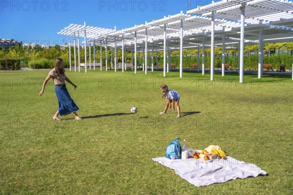 Mother and daughter playing soccer in a sunny green park, enjoying a picnic with blanket and
