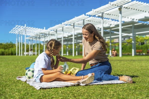 Mother and daughter enjoying a sunny summer picnic on green grass, sharing food and snacks while