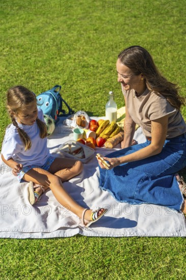 Mother and daughter share food and laughter on a picnic blanket in green grass, enjoying sunny park