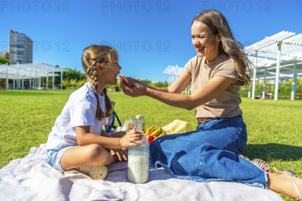 Young mother feeding her little daughter a sandwich during a sunny summer picnic on a blanket in