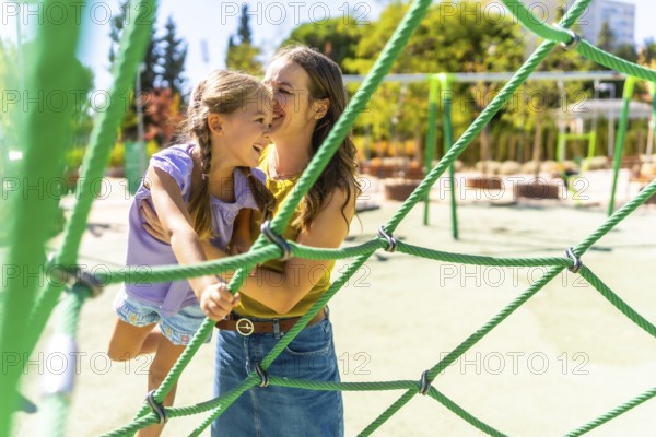 Mother supporting her happy young daughter climbing on a green rope playground structure, sharing a