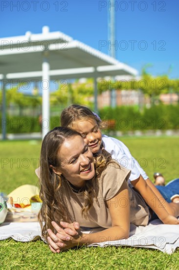 Mother and daughter lying together on a blanket, smiling and hugging, enjoying summer day outdoors