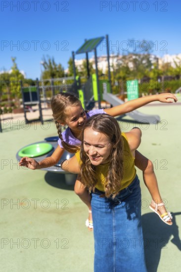 Mother gives daughter a piggyback at a sunny playground, both laughing and smiling as they enjoy
